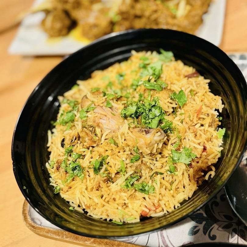 A black bowl filled with chicken biryani, garnished with fresh green cilantro, on a patterned napkin.