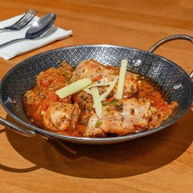 Chicken karahi, garnished with ginger, in a decorative metal serving dish on a wooden table.