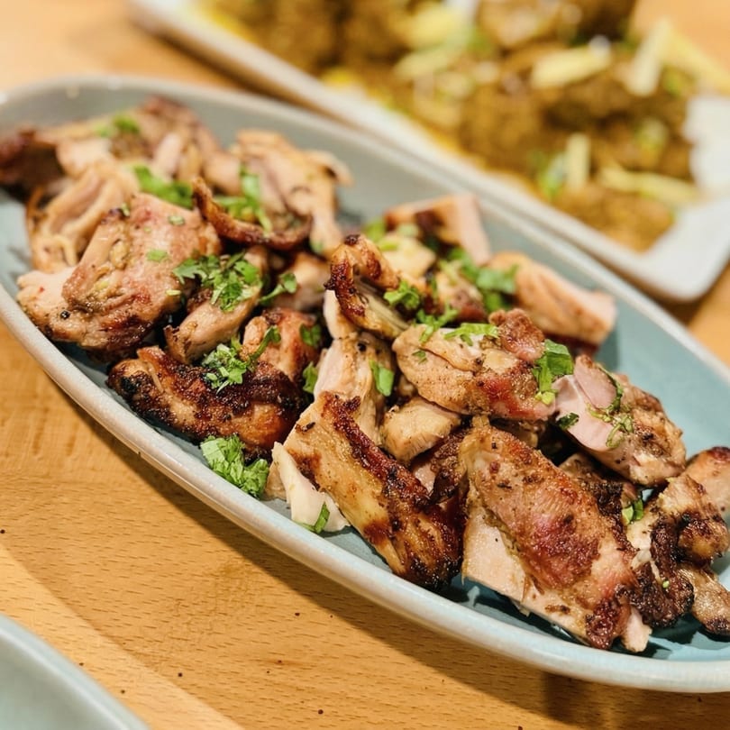 Grilled chicken pieces garnished with fresh herbs on a light blue platter, with another dish blurred in the background.