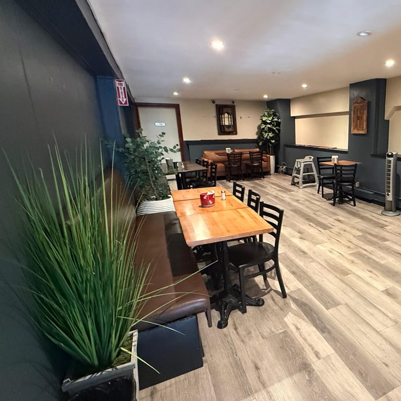 Restaurant interior with wooden tables, black chairs, a brown booth, and potted plants on a light wood floor.