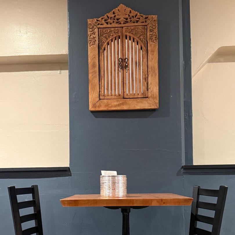 A small wooden table with two chairs and a tissue holder, against a blue wall with a carved wooden decoration.